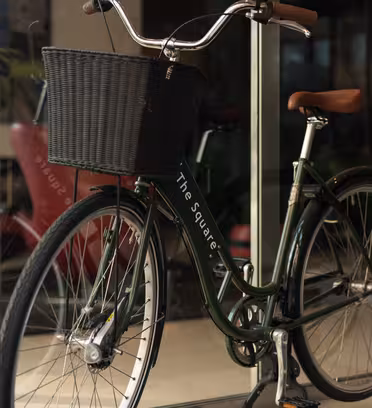 En grønn sykkel i vintagestil med en svart flettet kurv er parkert innendørs i nærheten av en glassdør. Scenen gjenspeiler et moderne urbant miljø med uskarpe utendørselementer synlig gjennom glasset.
