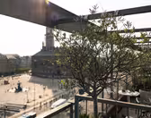 Utsikt fra en balkong med utsikt over et bytorg, med et lite tre i forgrunnen. Torget har historiske bygninger og en fontene. Bildet er opplyst av sollys, noe som gir det en varm tone.