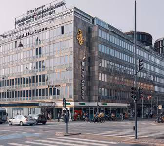 Et travelt bykryss med en moderne hotellbygning, «Hotel The Square», med fremtredende etiketter. Gaten er travel med biler, syklister og fotgjengere. Bygninger omgir området under klar himmel.