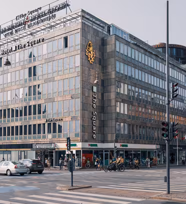 Et travelt bykryss med en moderne hotellbygning, «Hotel The Square», med fremtredende etiketter. Gaten er travel med biler, syklister og fotgjengere. Bygninger omgir området under klar himmel.