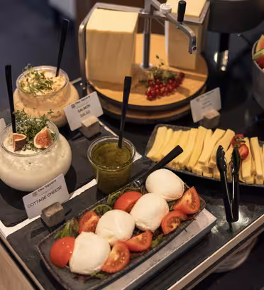 Et utvalg av matvarer inkludert skåler med cottage cheese, eggesalat, fløte med urter og pesto. Det er et fat med mozzarella og tomater, ost i skiver og en bolle med vannmelon. Friske bær legges ved siden av osten.