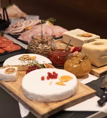 Et buffébord med forskjellige oster, oppskåret kjøtt, nøtter, frukt og krukker med syltetøy. Arrangementet er på treplater med kniver ved siden av, og settingen er elegant med fokus på variasjonen av smaker og teksturer.