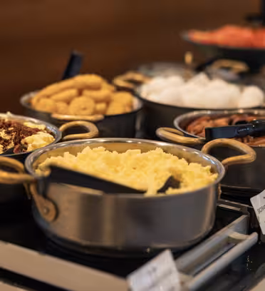 En frokostbuffé med ulike retter, inkludert eggerøre, pølser og smultringer. Maten stilles ut i metallgryter med serveringsutstyr på en disk. Små etiketter er synlige nær hver tallerken.