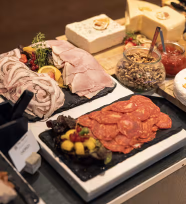 En selvbetjent delikatessedisk med assortert kjøtt og ost. Ulike oppskåret kjøtt, inkludert skinke og salami, vises på svarte brett. Store osteblokker og kiler, sammen med små krukker med krydder, fullfører oppsettet.