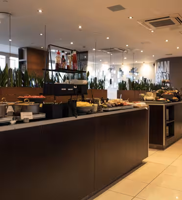 Et romslig bufféområde i en moderne restaurant med en rekke matvarer utstilt på mørke tredisker. Innstillingen er godt opplyst, med taklys og store vinduer. Grønne planter skiller buffeten fra sittegruppen i bakgrunnen.