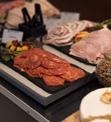 En delikatessedisk med pålegg, inkludert salami og skinke, arrangert på skiferheller. Tilbehør som blandede oliven og syltede grønnsaker serveres ved siden av, sammen med en bolle med nøtter. I bakgrunnen kan du se ostehjul og etiketter.