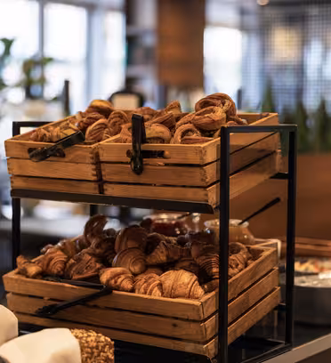 Trekasser fylt med nybakte croissanter er stablet på et metallstativ. Bakgrunnen viser en uskarp visning av en koselig kafé med planter og ekstra bakevarer.