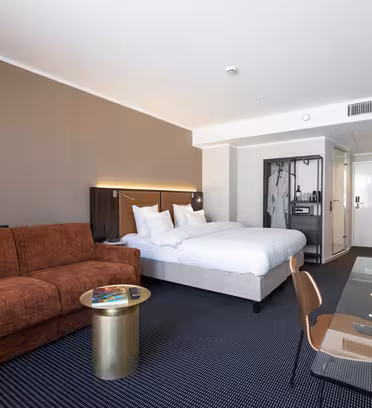 Moderne hotellrom med en stor seng, en brun sofa, et rundt gullfarget sidebord, et skrivebord med en stol, en veggmontert TV og en åpen garderobe. Rommet er innredet i nøytrale farger, har teppebelagt gulv og har et minimalistisk design.