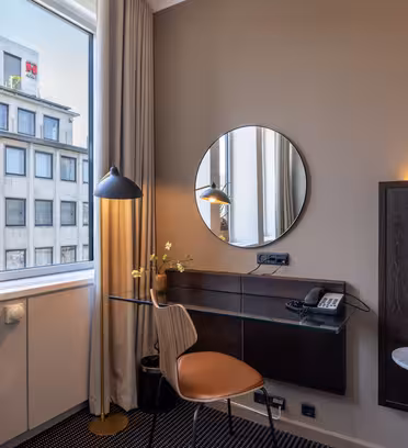 Et moderne hotellrom har et skrivebord med et rundt speil, en brun stol, en skrivebordslampe, en telefon og en vase med blomster. Store vinduer slipper inn naturlig lys og gir utsikt over en bygning utenfor.