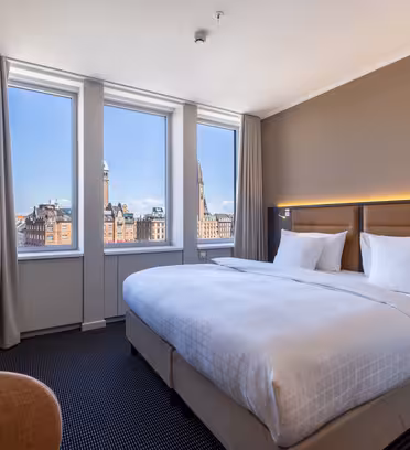 Moderne hotellrom med en stor, pent oppredd seng, hvitt sengetøy og dempet belysning. Tre høye vinduer viser et bybilde med historiske bygninger og blå himmel. Beige vegger og gardiner skaper en koselig og innbydende atmosfære.