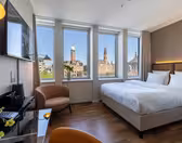Et moderne hotellrom med en stor seng, glasspult, lenestol og veggmontert TV. Tre høye vinduer gir utsikt over byen med historiske bygninger og kirkespir under en klar blå himmel.