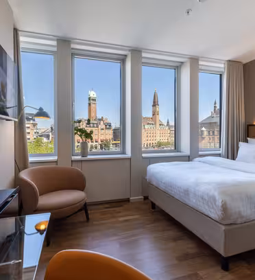 Et moderne hotellrom med en stor seng, glasspult, lenestol og veggmontert TV. Tre høye vinduer gir utsikt over byen med historiske bygninger og kirkespir under en klar blå himmel.