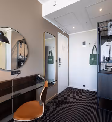 Moderne hotellrom med skrivebord, rundt speil, svart lampe, brun stol, stort veggspeil, skap med vannkoker, kopper og flasker, og en grønn veske hengende på døren. Innredningen er minimalistisk med nøytrale farger.