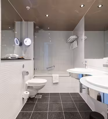 Moderne hotellbad med hvite flislagte vegger, svart flislagt gulv, to runde glassvasker med speil, vegghengt toalett, håndklestativ og badekar med glassvegg og montert dusjhode.