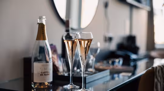 To fylte champagneglass og en åpen flaske musserende vin står på et glassbord i et stilig rom, med uskarpe bakgrunnselementer, inkludert et speil og diverse gjenstander.