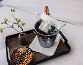 På et brett på en seng står en vase med grønne blomster, en skål med forskjellige nøtter og en metallisbøtte som inneholder en champagneflaske, to glass og en brettet hvit serviett.