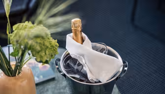 En flaske champagne pakket inn i en hvit serviett står i en isbøtte ved siden av to tomme glass, med en grønn plante i en vase i nærheten på et bord.