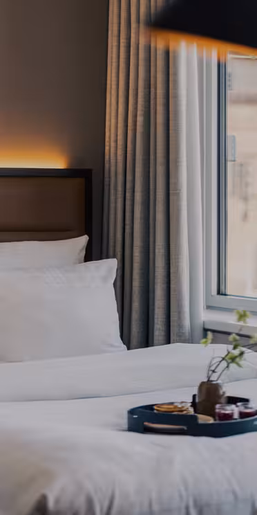 En pent oppredd hotellseng med hvitt sengetøy speiler seg i et rundt speil. På sengen er det et frokostbrett. Store vinduer med gardiner slipper inn naturlig lys, og en vase med grønne planter står i vinduskarmen.
