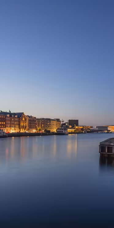 Skumringsutsikt over et rolig bybilde ved vannet med fargerike bygninger langs elven. Gatelys lyser varmt og reflekteres i det rolige vannet, mens himmelen går fra blått til dypere skumring.