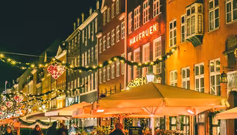 Festlig gatebilde om natten med opplyste hjerteformede dekorasjoner som henger over en brosteinsbelagt sti med historiske bygninger med flere etasjer. Diners sitter under store parasoller utenfor en varmt opplyst restaurant. Folk går og nyter den muntre atmosfæren.