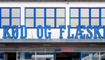 Folk sitter på fluktstoler foran en bygning med store blå vinduer og den blå inskripsjonen «KØD OG FLÆSK». Over inngangen henger en dyreskulptur, trolig en gris eller en ku. Det er en solrik dag med en livlig atmosfære.