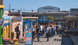 En yrende utendørs markedsscene med folk samlet i nærheten av fargerike matboder. Et stort industribygg med "Refshaleøen" skrevet på står i bakgrunnen. Stringlys henges over hodet, og skaper en livlig atmosfære under klar himmel.