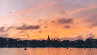 En rolig solnedgang over en rolig innsjø, med vann som reflekterer himmelens livlige farger. Silhouettede bygninger og trær langs den fjerne kysten, mens fugler flyr over den rosa og oransje himmelen.