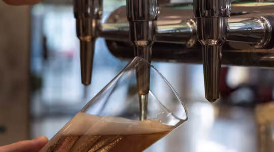 En person heller øl i et skrått, høyt glass fra en skinnende krom ølkran. Skummet dannes på toppen av den gule væsken. Bakgrunnen er uskarp, med flere trykk og en subtil innendørs setting.