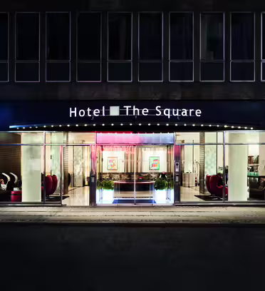 Bildet viser den opplyste inngangen til Hotel The Square om natten. Fasaden er i glass med synlige sittegrupper innvendig. Hotellnavnet vises tydelig over inngangen.