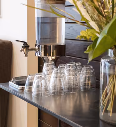 En vanndispenser på et metallbord med en stabel plastkopper til høyre. En stor glassvase med grønne og hvite blomster står i forgrunnen. En brun sofa og veggkunst er synlig i bakgrunnen.