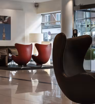 Moderne hotellobby med elegante, mørke svingstoler og røde eggestoler rundt et lavt bord. En stor fotokollasje henger på veggen. Glassdører avslører en gateutsikt utenfor. Lys og innbydende plass med blanke fliser på gulv.