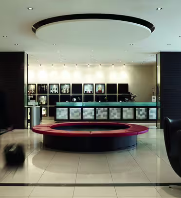 En moderne hotellobby med en sirkulær rød sittegruppe i sentrum. En person i dress går raskt med en koffert. Resepsjonen er i bakgrunnen, flankert av hyller og dekorativ belysning, som skaper en elegant, profesjonell atmosfære.