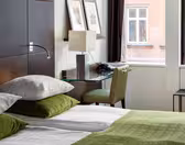 Et hotellrom med en pent oppredd seng med grønt og hvitt sengetøy. Et skrivebord med glassplate har en lampe, telefon og innrammede bilder. To store vinduer med mørke gardiner gir utsikt til en murbygning utenfor.