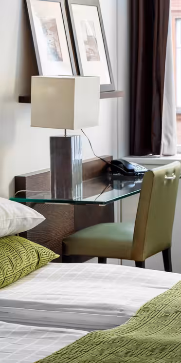 Et hotellrom med en pent oppredd seng med grønt og hvitt sengetøy. Et skrivebord med glassplate har en lampe, telefon og innrammede bilder. To store vinduer med mørke gardiner gir utsikt til en murbygning utenfor.