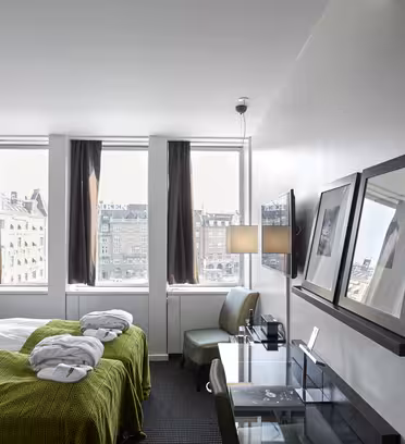 Moderne hotellrom med to pent oppredde enkeltsenger med grønne plagg. En veggmontert TV, skrivebord og stol er til høyre. Store vinduer med mørke gardiner gir utsikt over byen. Veggene er hvite, og rommet har en ren, minimalistisk design.