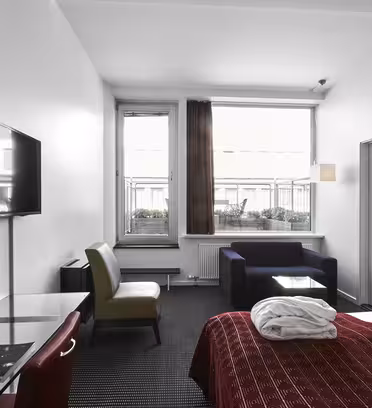 Moderne hotellrom med en stor seng med rødt sengetøy og en sammenbrettet badekåpe. Det er en veggmontert TV, et skrivebord med stol og et sittehjørne med sofa. Stort vindu med gjennomsiktige og blendingsgardiner gir naturlig lys.