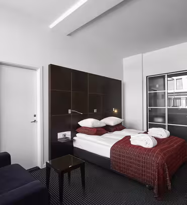 Moderne hotellrom med dobbeltseng utsmykket med rødmønstret plagg og puter. En hvit badekåpe er plassert på sengen. Rommet har en mørk sofa, et salongbord i glass og en garderobe med skyvedører. Veggene er hvite og teppet er mørkt.