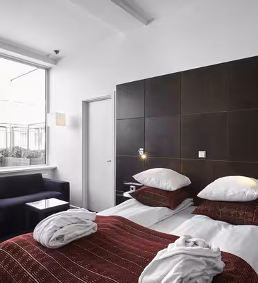 Et moderne hotellrom med to pent oppredde enkeltsenger med hvitt og rødt sengetøy. Et stort vindu slipper inn naturlig lys, og avslører en mørk sofa og et lite glassbord. Rommet er innredet i en minimalistisk stil med mørkt trepanel.