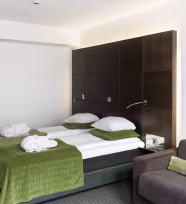 Hotellrom med dobbeltseng med grønt sengetøy og to badekåper. En mørkebrun sengegavl har festet leselys. Det er en grå sofa med en matchende grønn pute, og en hvit dør fører til inngangen. Rommet har moderne innredning.
