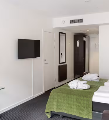 Et hotellrom med dobbeltseng dekket av grønt og hvitt sengetøy. To badekåper er pent plassert på sengen. Rommet har veggmontert TV, skrivebord i glass med lampe og garderobeskap. Veggene er hvite, og gulvet har mørkt teppe.