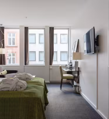 Moderne hotellrom med to senger dekket av grønne tepper og badekåper lagt på toppen, en flatskjerm-TV montert på en hvit vegg, et skrivebord med lampe og stol, og store vinduer med utsikt over nabobygningene.