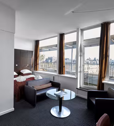Moderne hotellrom med store vinduer med utsikt over byen. Rommet har en seng med hvitt sengetøy, en sofa, et glasssalongbord og svarte stoler. Innredningen er minimalistisk med nøytrale toner og naturlig lys som fyller rommet.