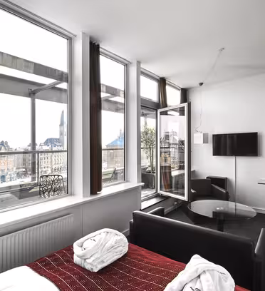 Et moderne hotellrom med store vinduer gir utsikt over byen. Rommet har en seng, et rødt teppe, en svart sofa, et lite bord, veggmontert TV og kunstverk. En dør åpner ut til en balkong. Bybildet har historiske bygninger og et klokketårn.