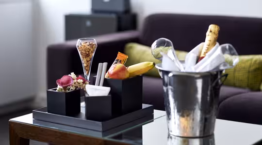 En moderne stue med et glassbord med frukt og snacks, inkludert bananer, et eple, nøtter og sjokolade. En avkjølt flaske champagne i en isbøtte står ved siden av arrangementet. En mørk sofa med grønne puter er i bakgrunnen.