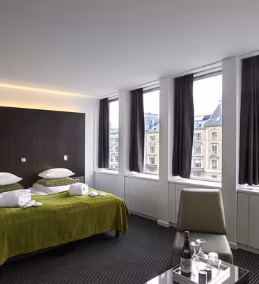 Et moderne hotellrom har en dobbeltseng med et grønt innslag og to hvite badekåper. Tre store vinduer avslører et bybilde med historiske bygninger. Rommet inkluderer en grønn stol og et bord med flaskevann og glass.