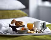 Et frokostbrett på en seng med et grønt teppe. Det inkluderer croissanter i en kurv, et kokt egg, en bolle med tørkede aprikoser, ost, salami, agurkskiver, et glass appelsinjuice, et hvitt krus og bestikk.