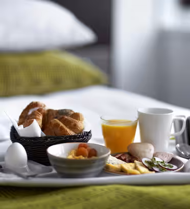Et frokostbrett på en seng med et grønt teppe. Det inkluderer croissanter i en kurv, et kokt egg, en bolle med tørkede aprikoser, ost, salami, agurkskiver, et glass appelsinjuice, et hvitt krus og bestikk.