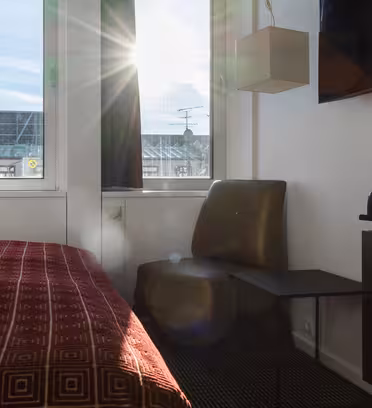 Sollyset strømmer inn i et hotellrom gjennom et vindu, og lyser opp en stol og et sidebord. En seng med et mønstret rødt deksel er i forgrunnen. En TV og et brett med vannkoker og kopper vises til høyre.