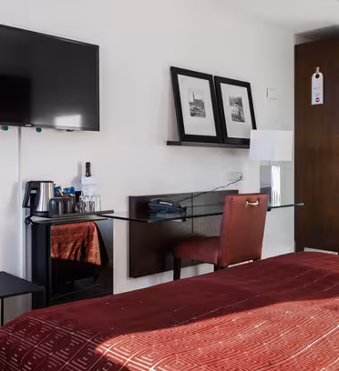 Moderne hotellrominteriør med veggmontert TV, et lite skrivebord med en rød stol, to innrammede bilder og en lampe. Sengen er dekket med et rødt teppe med et geometrisk mønster. Et lite bord rommer en vannkoker og gratis ting.