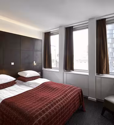 Hotellrom med dobbeltseng dekket av rødmønstret sengetøy, satt mot en hodegjerde i mørkt tre. Det er tre vinduer med brune gardiner, en stol i nærheten og en urban utsikt utenfor med en moderne glassbygning.